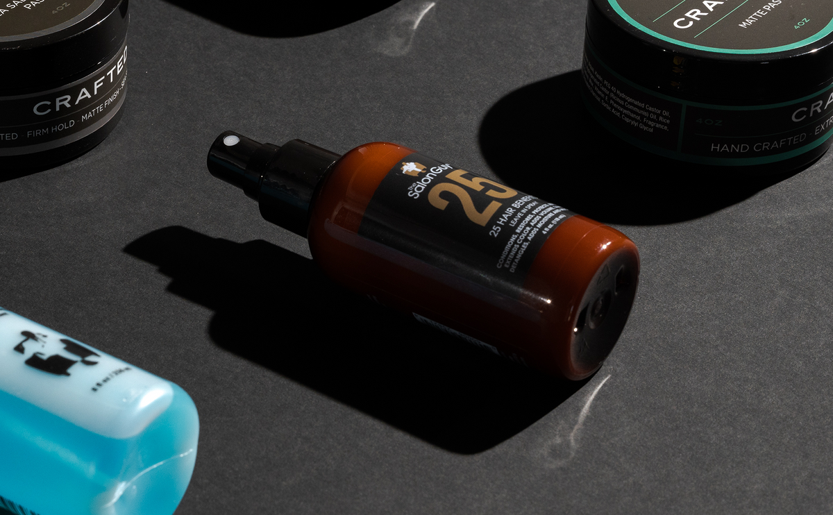 TheSalonGuy 25 Leave-In Treatment Spray beside styling clay and paste on a dark studio surface